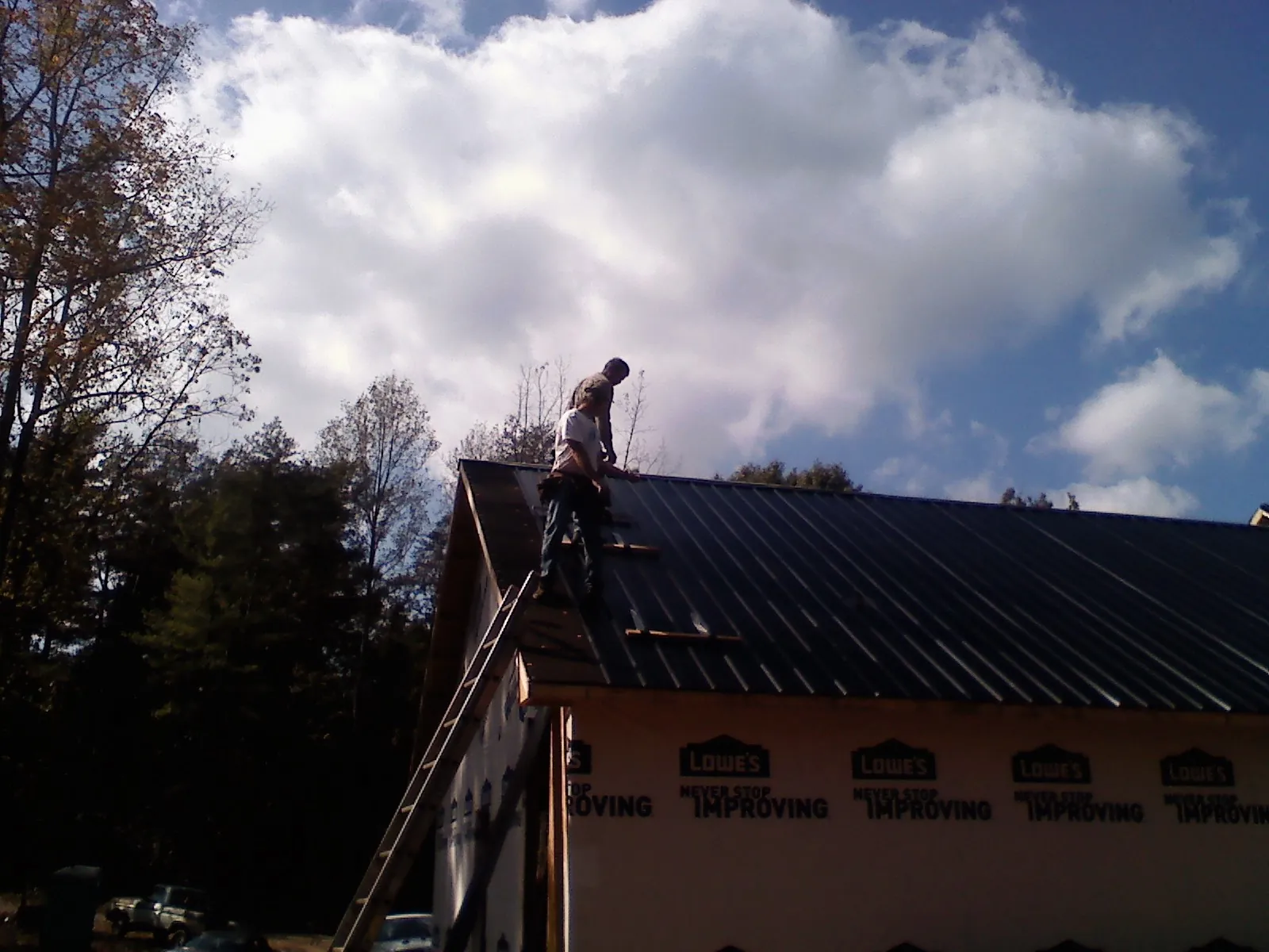 Roofer fastening dark metal panels high against a cloudy sky, from a Roof Inspection job in Groton